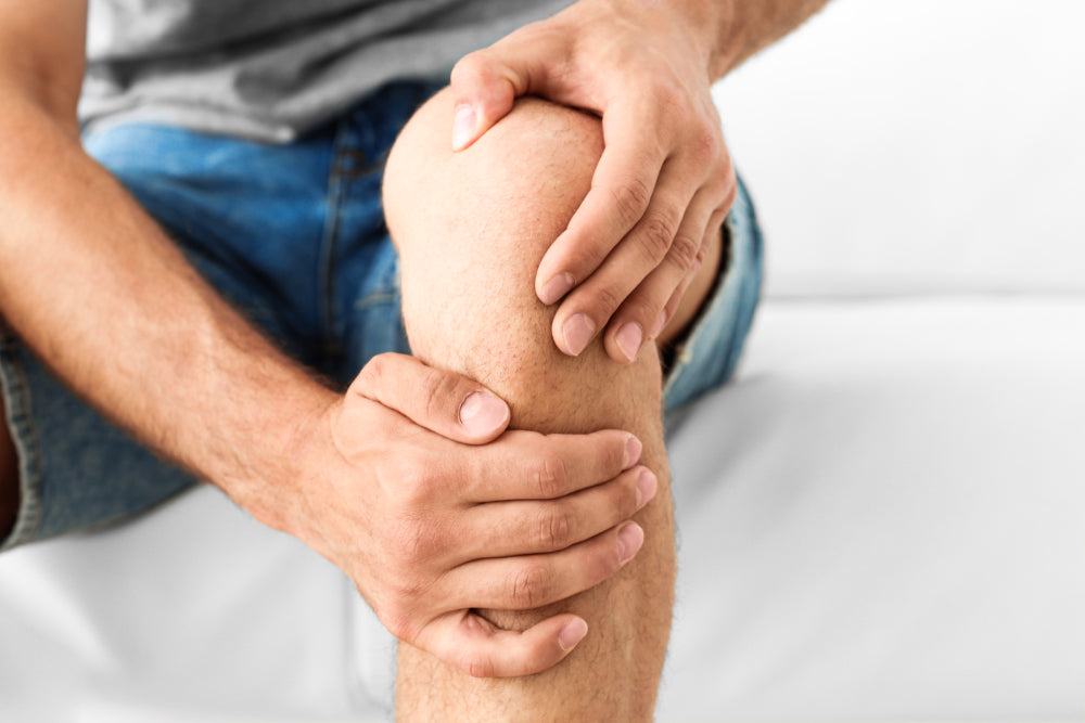 Nahaufnahme eines Knies, das von zwei Händen gehalten wird, mit sichtbarem Hautdetail und Jeans im Hintergrund