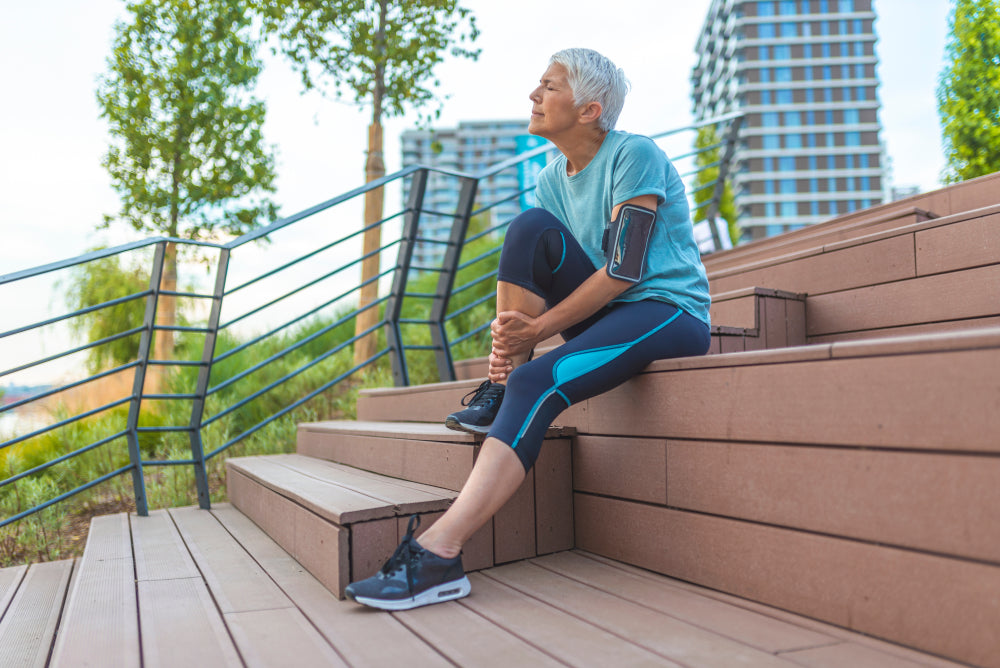 Ältere Frau in blauem Sportoutfit sitzt auf Holzstufen und hält ihr rechtes Sprunggelenk.
