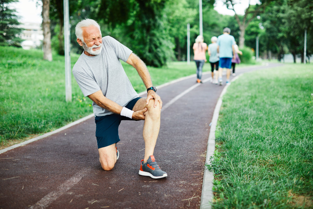 Älterer Mann in Sportkleidung hält sich schmerzend das rechte Knie auf einem Parkweg aus Pflastersteinen.