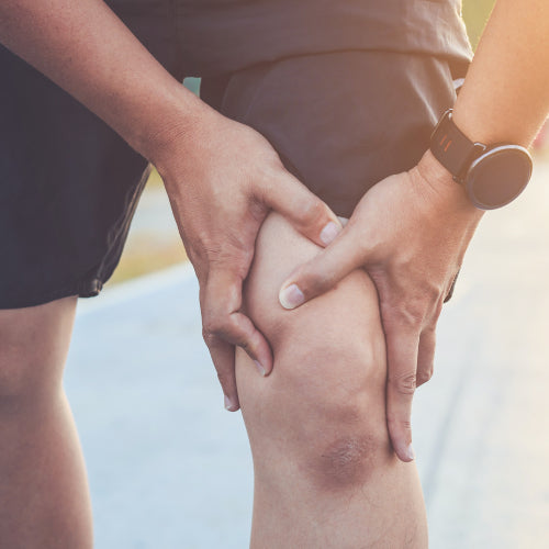Nahaufnahme einer Person in schwarzen Shorts, die das rechte Knie hält, auf einem gepflasterten Weg im Freien.