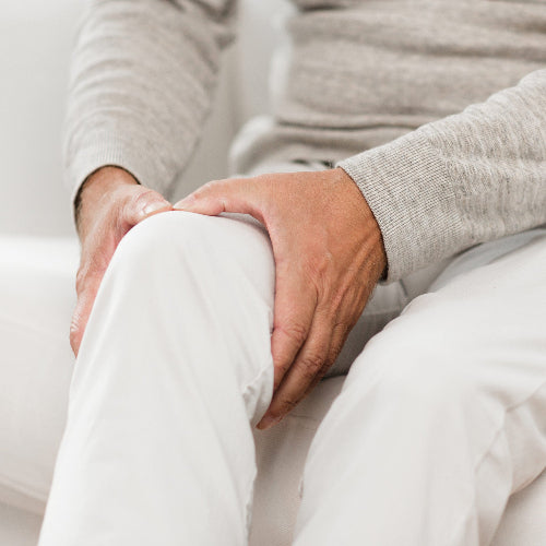 Hände halten schützend ein schmerzendes Knie, bekleidet mit weißen Hosen und einem grauen Pullover.