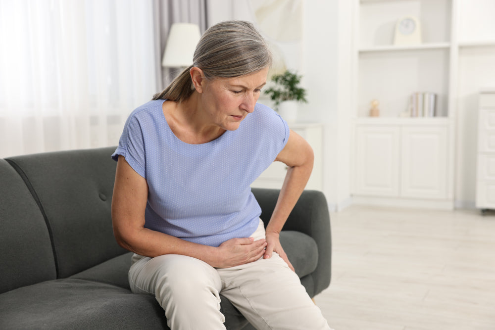 Ältere Frau hält schmerzerfüllt ihre Hüfte und ihren Bauch im Wohnzimmer.