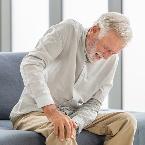Älterer Mann hält schmerzhaft sein rechtes Knie auf grauem Sofa in modernem, lichtdurchflutetem Raum sitzend.